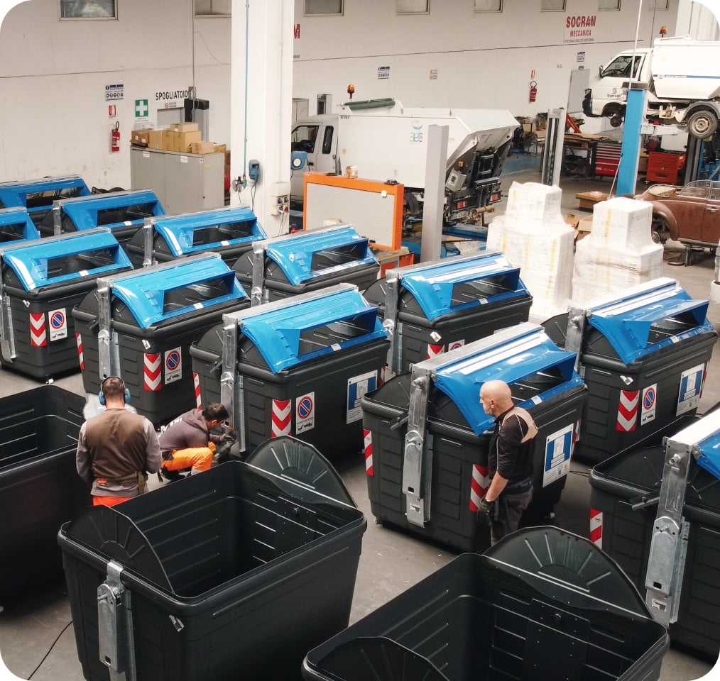 Veduta dall'alto del capannone Socram Meccanica con numerosi cassonetti stradali neri a coperchio blu in fase di allestimento, con tre operai al lavoro tra i contenitori.