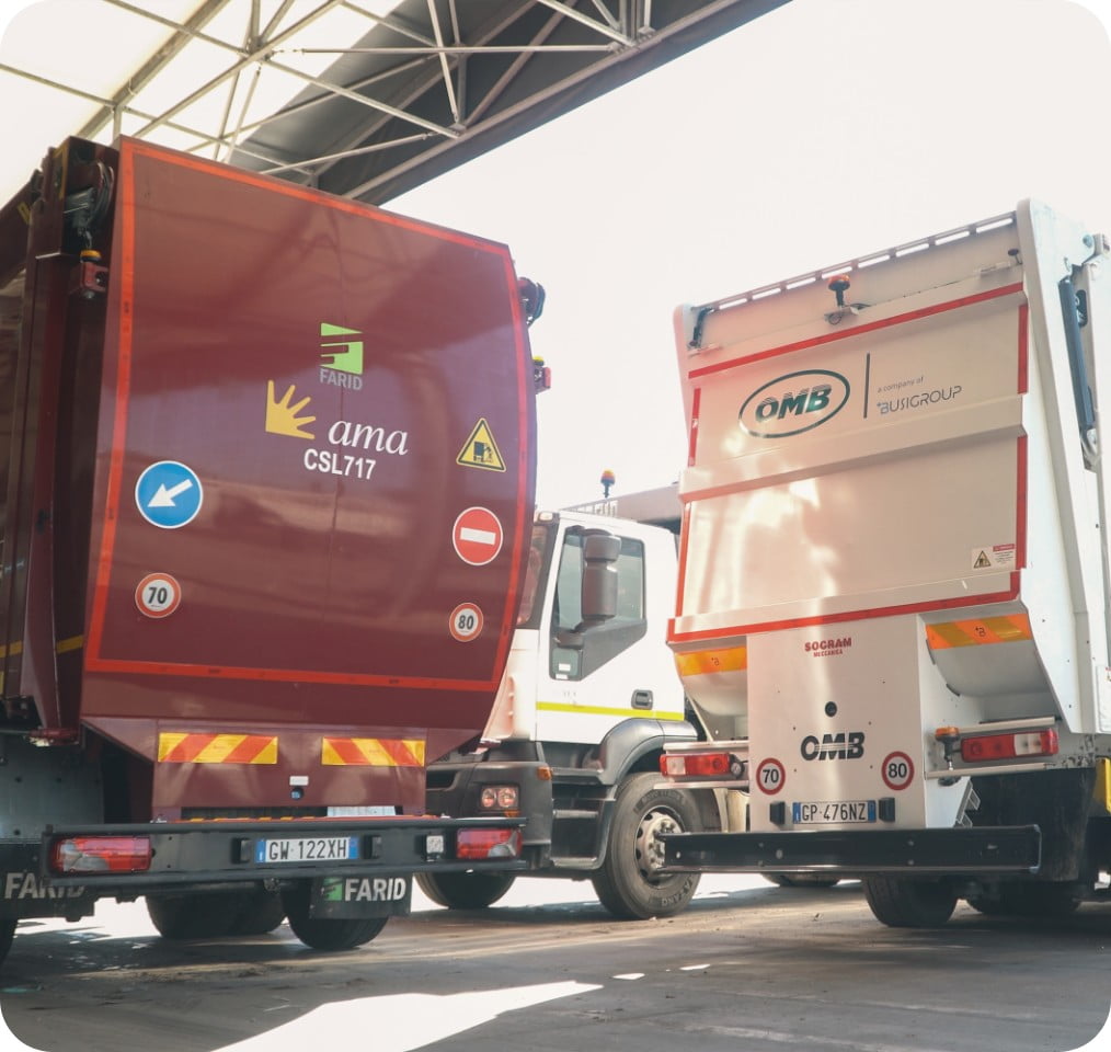 Vista posteriore di due autocompattatori affiancati sotto una tettoia: a sinistra un mezzo bordeaux AMA Roma FARID identificativo CSL717, a destra uno bianco con vasca OMB Busi Group con logo Socram Meccanica.