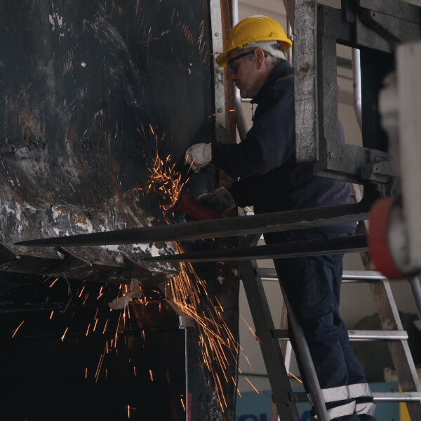 Operaio con casco giallo e tuta blu in piedi su una scala che esegue una molatura su una grande struttura metallica scura, con scintille arancioni visibili nell'officina.