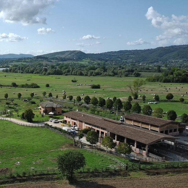 Veduta aerea di un'azienda agricola con edifici rurali in mattoni e tetto in tegole, immersa in una vasta pianura verde con rotoballe di fieno sparse nei campi, colline boscose e cielo parzialmente nuvoloso sullo sfondo.