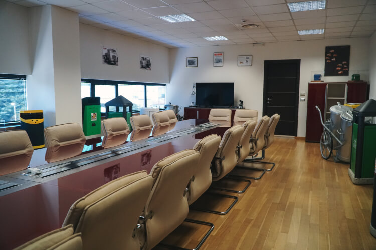 Sala riunioni aziendale con lungo tavolo bordeaux, sedie in pelle beige, fotografie storiche di veicoli alle pareti e, in primo piano, cassonetti per la raccolta differenziata utilizzati come elementi d'arredo.