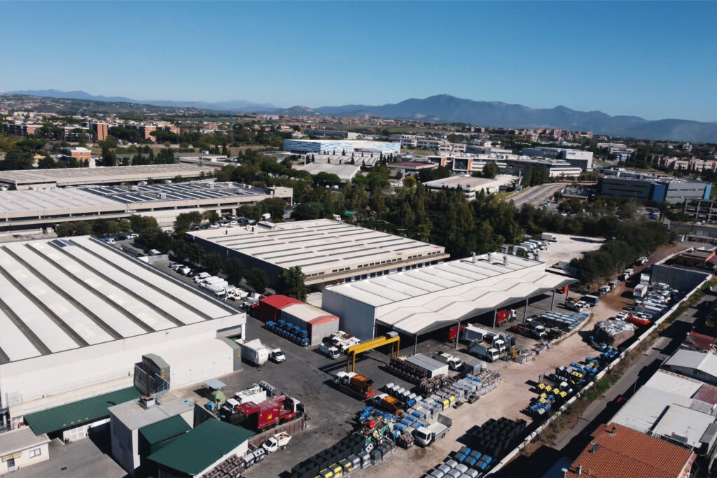 Veduta aerea del polo industriale Fantauzzi con capannoni, piazzali con autocompattatori, cassonetti e attrezzature per l'igiene urbana, circondato da un'area urbana e con le montagne dei Castelli Romani sullo sfondo.