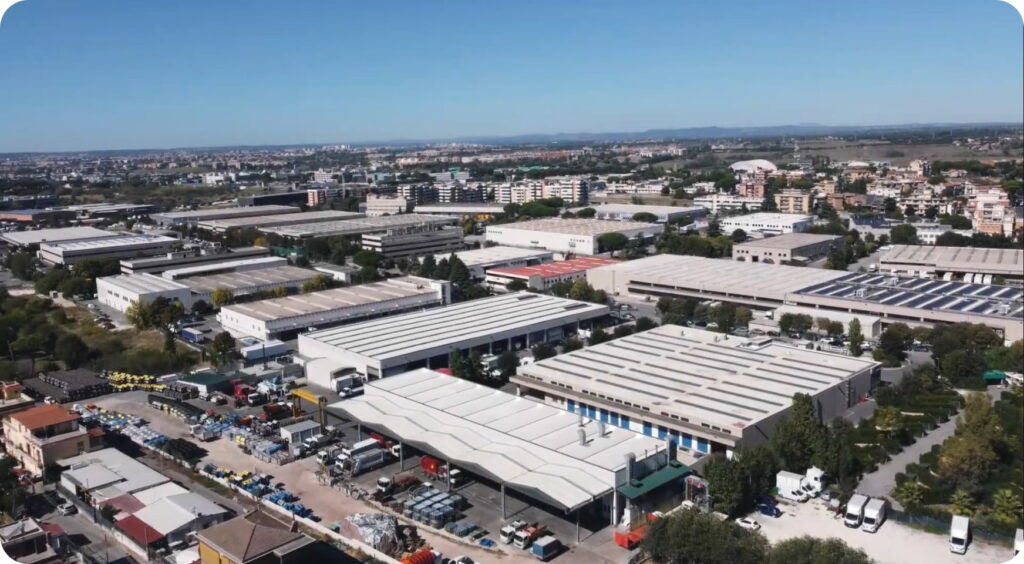 Veduta aerea di un ampio polo industriale con capannoni, piazzali con veicoli e attrezzature per la raccolta rifiuti, circondato da un'area urbana con edifici residenziali e commerciali, ripreso in una giornata di sole con cielo blu.