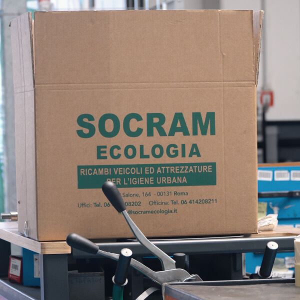 Scatola di cartone con logo Socram Ecologia e dicitura "Ricambi veicoli ed attrezzature per l'igiene urbana" posizionata su un transpallet in un magazzino.