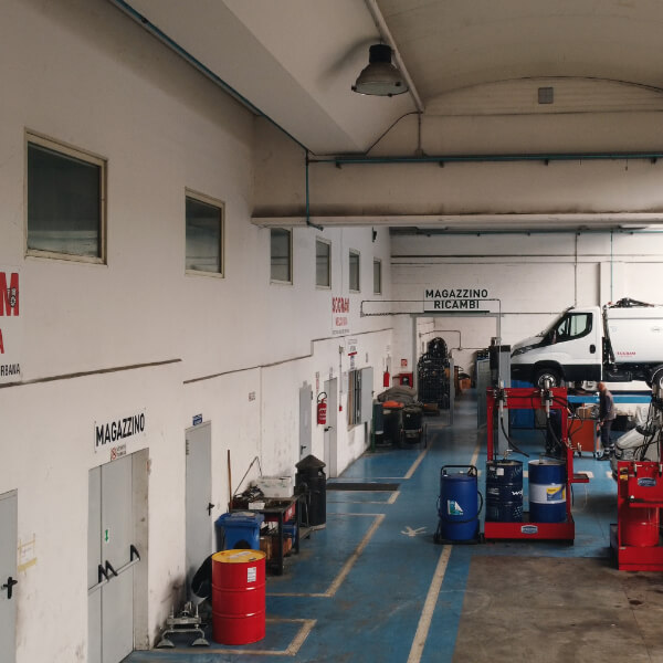 Corridoio interno dell'officina Socram Meccanica con magazzino ricambi, fusti industriali rossi e blu, attrezzature per pneumatici e un furgone bianco sul ponte sollevatore sullo sfondo.