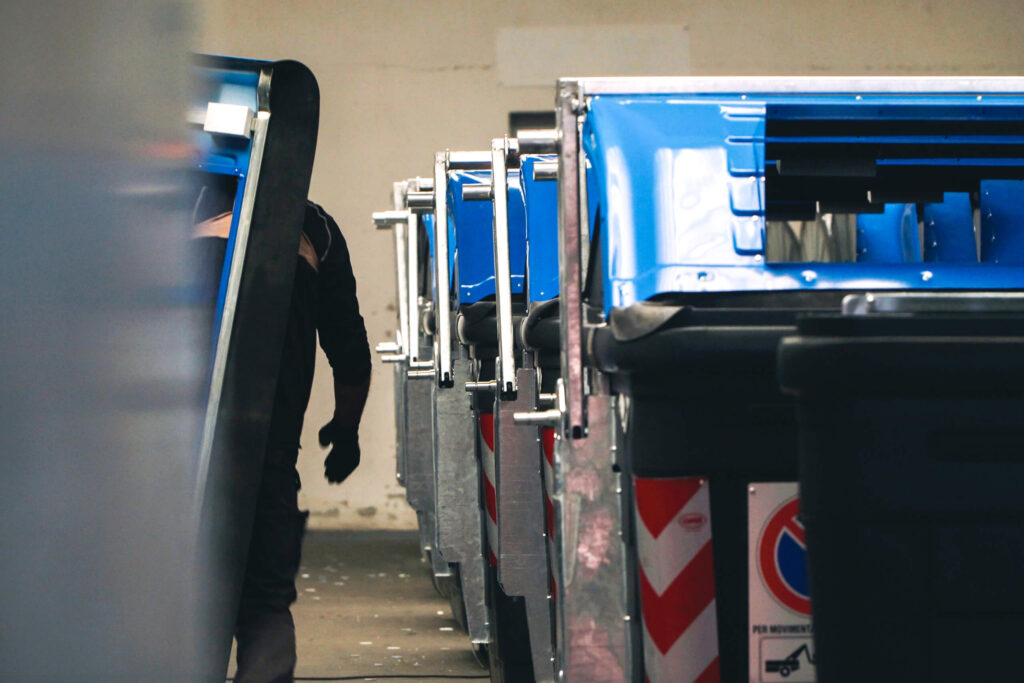 Operaio in tuta nera che lavora tra una fila di cassonetti stradali neri con coperchi blu e barre di aggancio in acciaio, ripresi in prospettiva ravvicinata in un magazzino.