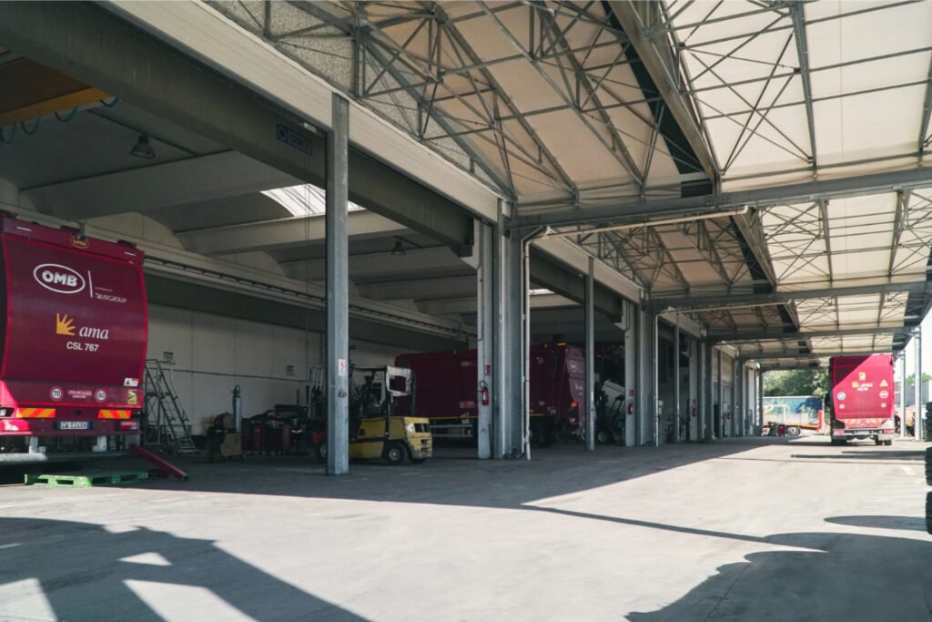 Interno di un ampio capannone industriale con più baie di officina contenenti autocompattatori bordeaux AMA Roma con logo OMB, un carrello elevatore giallo e attrezzature di servizio, ripreso in prospettiva lungo la navata centrale.