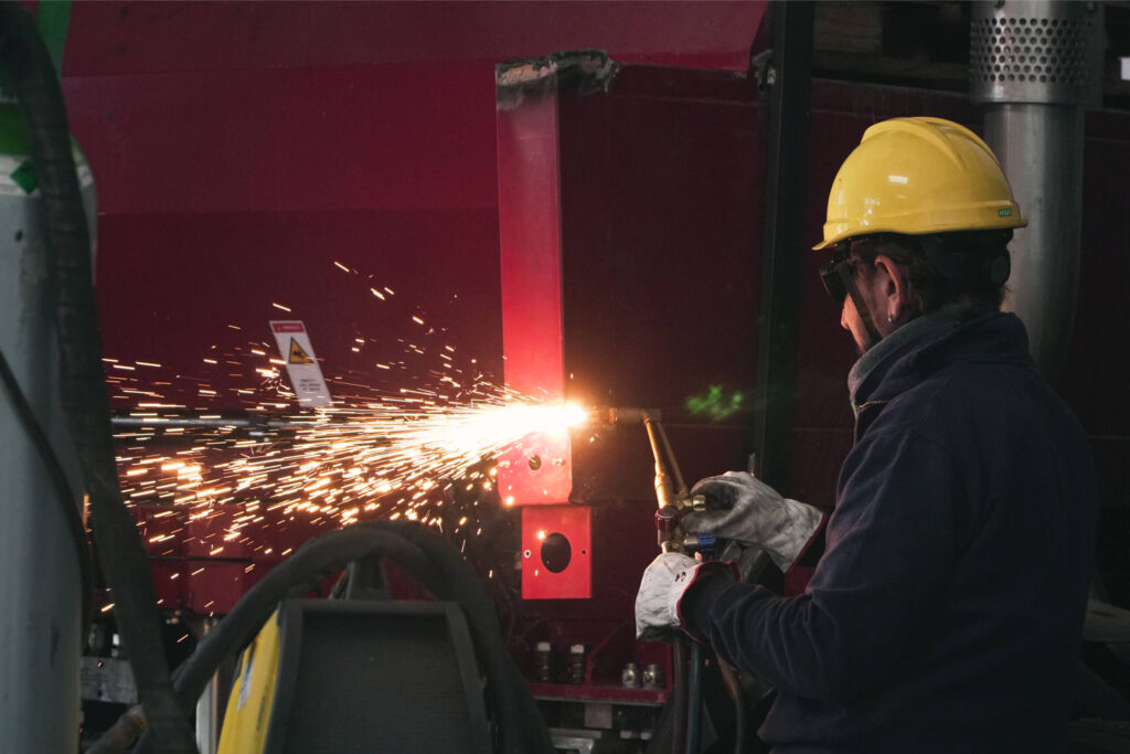 Operaio con casco giallo e guanti protettivi che esegue un taglio ossiacetilenico su una struttura metallica bordeaux di un autocompattatore, con una cascata di scintille arancioni ben visibile.