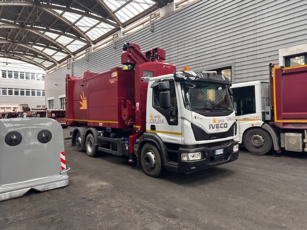 Autocompattatore IVECO bianco e bordeaux con sistema di caricamento aereo AMA Roma, identificativo CBIL014, parcheggiato in un capannone industriale con altri veicoli sullo sfondo.
