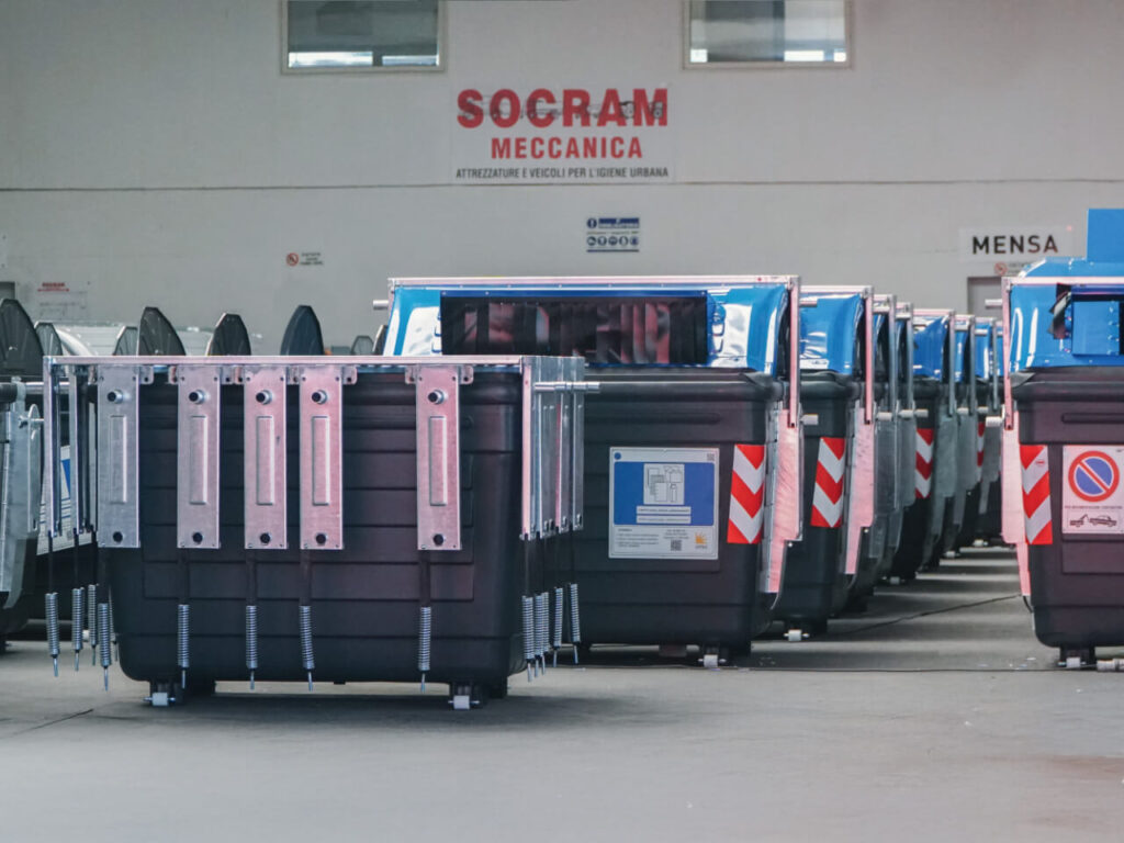 Fila di cassonetti stradali neri con coperchi blu e barre metalliche di aggancio, pronti per la consegna all'interno del magazzino Socram Meccanica, azienda specializzata in attrezzature e veicoli per l'igiene urbana.