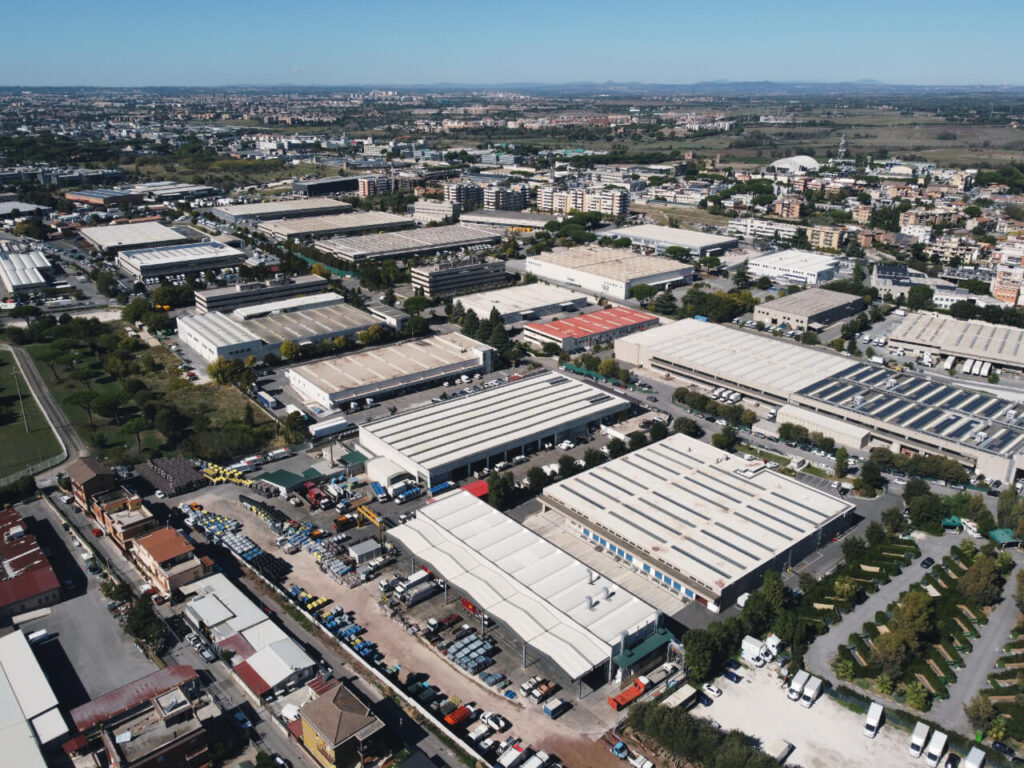Veduta aerea di un ampio polo industriale con capannoni, piazzali con veicoli e attrezzature per la raccolta rifiuti, circondato da un'area urbana con edifici residenziali e commerciali, ripreso in una giornata di sole con cielo blu.