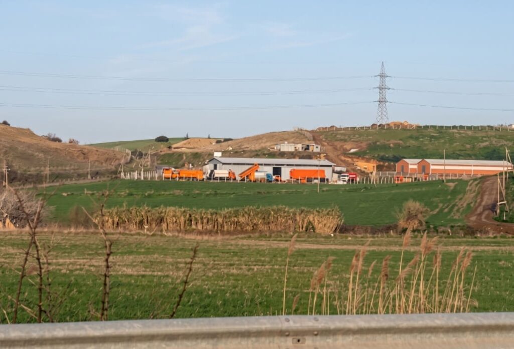 Veduta panoramica della sede storica di Socram con una fila di camion arancioni in attesa davanti a capannoni industriali, circondato da colline verdi.