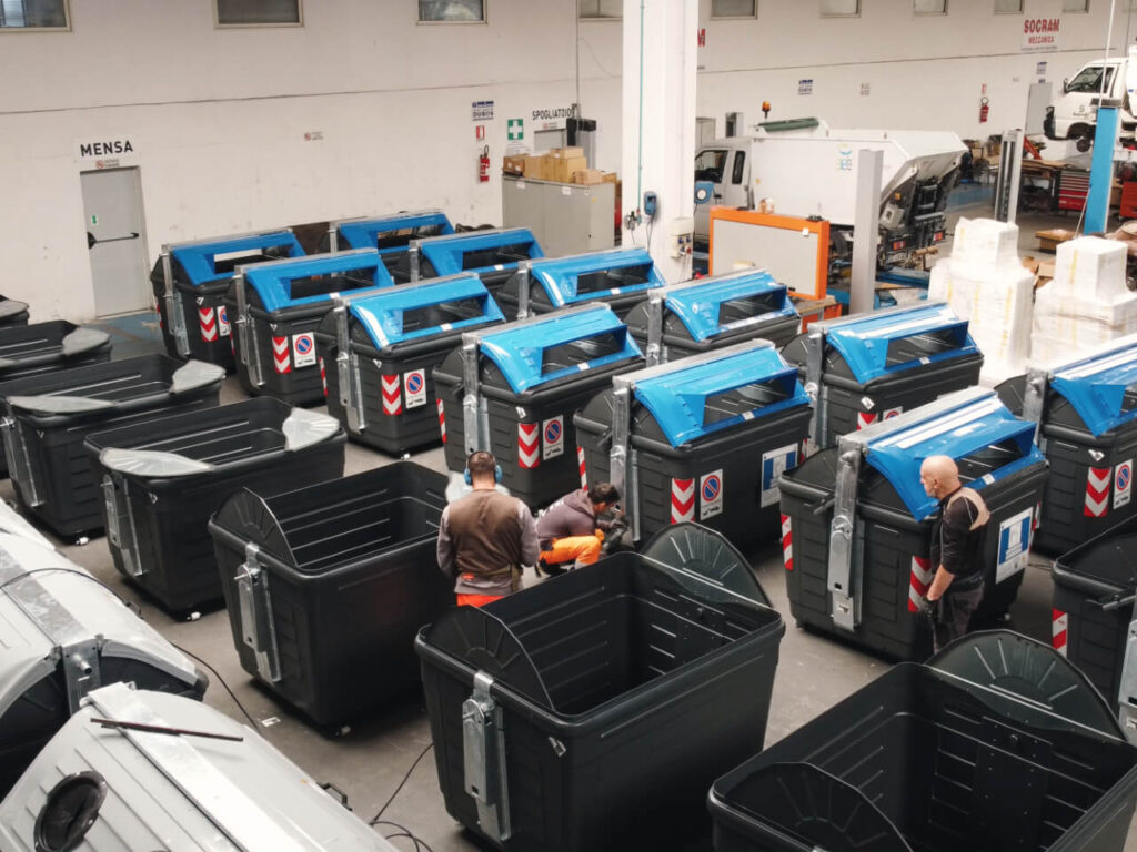 Veduta dall'alto del capannone Socram Meccanica con numerosi cassonetti stradali neri a coperchio blu in fase di allestimento, con tre operai al lavoro tra i contenitori.