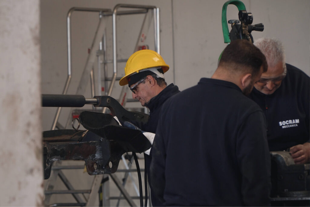 Due tecnici Socram Meccanica in tuta blu al lavoro su un componente meccanico: uno indossa casco giallo e occhiali protettivi, l'altro, con capelli grigi, esamina il pezzo da vicino.