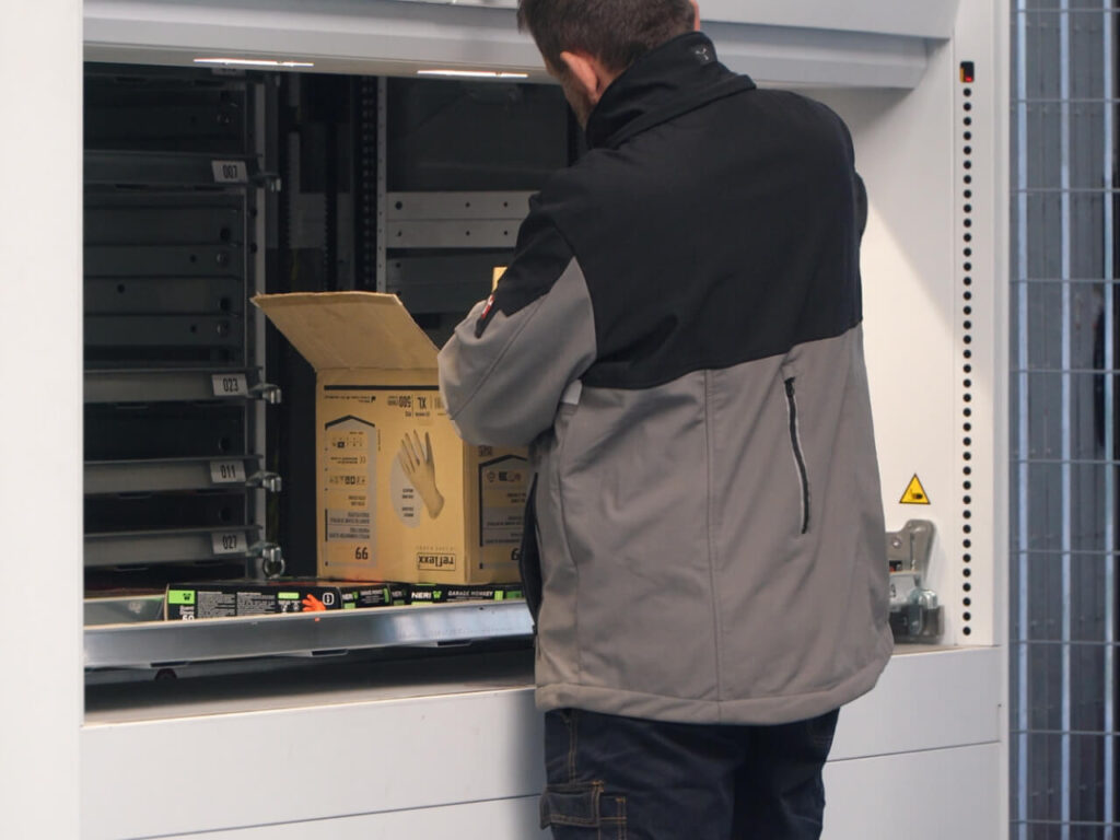 Operatore di magazzino che preleva una scatola di ricambi da un sistema di scaffalatura verticale automatizzata, ripreso di spalle in un magazzino ricambi.