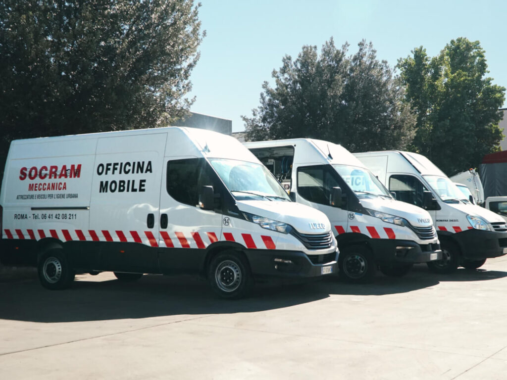 Fila di furgoni IVECO Daily bianchi con livrea a bande rosse e scritta "Socram Meccanica – Officina Mobile", parcheggiati in un piazzale in una giornata di sole.
