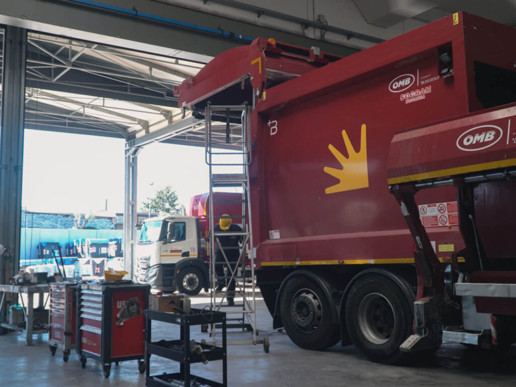 Autocompattatore bordeaux OMB Busi Group con logo Socram in manutenzione all'interno di un'officina, con carrello porta-utensili e scala a trabattello affiancati, e un secondo mezzo AMA visibile sullo sfondo attraverso il portone aperto.